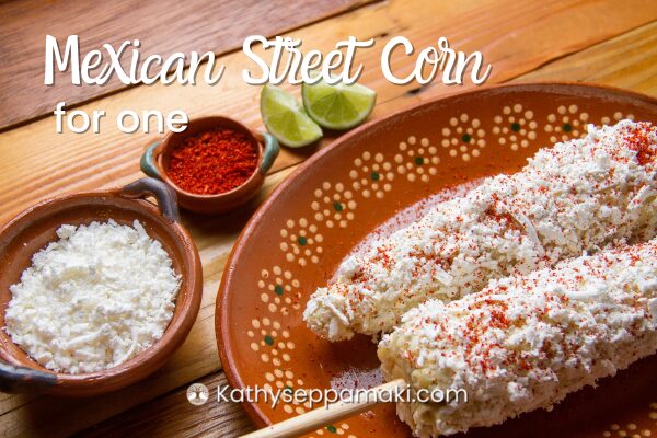 Mexican Street Corn for One recipe title with a picture of two ears of Mexican street corn on a wooden plate sitting on a table next to a small bowl of white cheese and a small bowl of paprika.