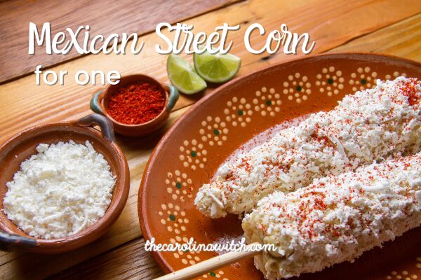 Mexican Street Corn for One recipe title with a picture of two ears of Mexican street corn on a wooden plate sitting on a table next to a small bowl of white cheese and a small bowl of paprika.