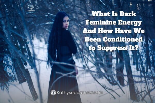 What Is Dark Feminine Energy and How Have We Been Conditioned to Suppress it? Blog post title with a picture of a woman standing on a forest path walking away from us, but turning around and looking back at us.